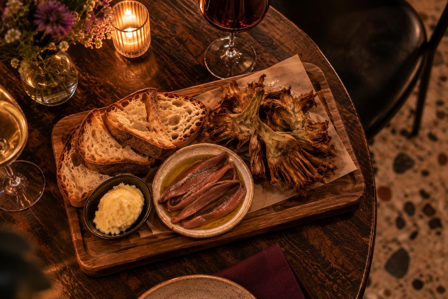 Un tagliere sul tavolino a lume di candela: pane tostato e burro salato, alici sott'olio e carciofi alla giudia, con un calice di rosso e uno di bianco